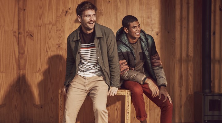 Two male models sitting on a wooden box wearing brown jackets and pants laughing.