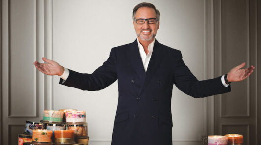 Founder of Slatkin + Co Harry Slatkin standing against a white wall with candles on both sides of him.