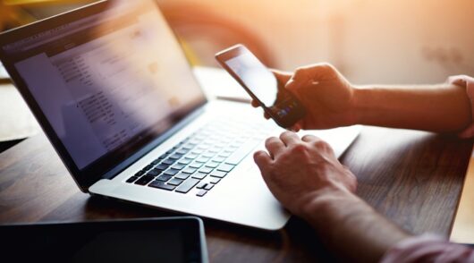 Image of person using a laptop and holding a phone.