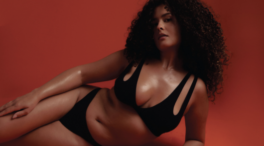 A curly-haired woman wearing a black set from the lingerie brand Nuudi System against a red background.