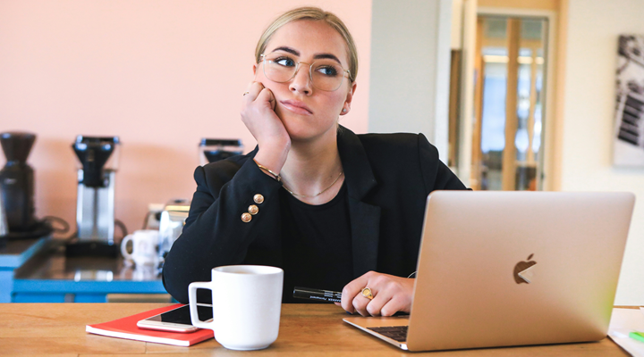 a young woman wearing a black blazer sitting at a laptop with a bored look on her face
